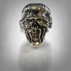 Pu&ograve; includere: Un ciondolo a forma di teschio in argento con un'espressione dettagliata a bocca aperta. Il teschio ha una superficie testurizzata e sembra essere fatto di metallo.