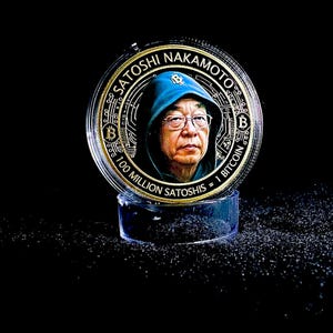 Puede incluir: Una moneda dorada con la imagen de Satoshi Nakamoto, el creador de Bitcoin, con una chaqueta con capucha azul y gafas. La moneda se exhibe sobre un soporte cilíndrico transparente sobre un fondo negro. El texto de la moneda dice "Satoshi Nakamoto" y "100 Millones de Satoshis = 1 Bitcoin."