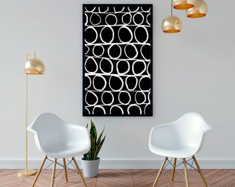 May include: Black and white abstract art print with a repeating pattern of connected circles. The artwork is framed in a black border and hangs on a white wall. The room features white chairs, a gold floor lamp, and gold pendant lights.