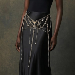 May include: A gold-toned body chain with dangling strands and decorative embellishments. The jewelry piece is worn over a black dress, creating a layered effect. The body chain features a central design with intricate details.