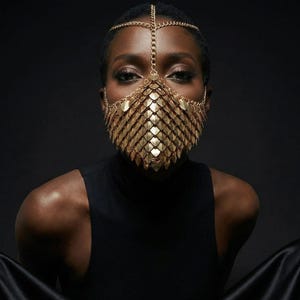 May include: A gold-colored face mask with a scale-like design, paired with a matching headpiece. The mask covers the nose and mouth, and the headpiece is connected by delicate chains. The person is wearing a black turtleneck and sitting against a dark background.
