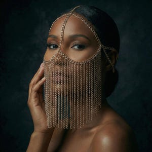 Puede incluir: Una máscara de cadena dorada con una diadema, que cubre los ojos y la parte inferior de la cara. La máscara está hecha de pequeñas cadenas conectadas que cuelgan. El fondo es una superficie oscura y texturizada.