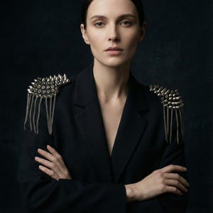 Pode incluir: Blazer preto com enfeites de ombro prateados e pontiagudos e franjas de corrente penduradas. A jaqueta é usada por uma pessoa com os braços cruzados, contra um fundo escuro. As ombreiras adicionam um estilo ousado e moderno.
