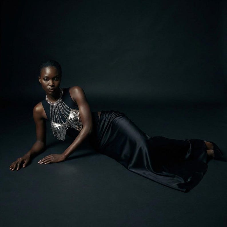 May include: A black sleeveless top with a silver chainmail design and a long black satin skirt. The model is lying on a dark surface, creating a dramatic and elegant look. The outfit is suitable for evening wear.