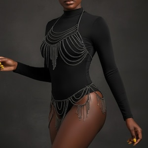 Op de afbeelding: Een zwarte bodysuit met een hoge halslijn en lange mouwen. De bodysuit is versierd met een gelaagd kettingharnas dat over de borst en taille drapeert, met bungelende kettingfranjes. De achtergrond is donkergrijs.
