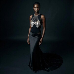 May include: A black sleeveless bodysuit with a silver chainmail top and a long black satin skirt. The skirt has a high waist and a slight train. The model is standing in front of a dark background.