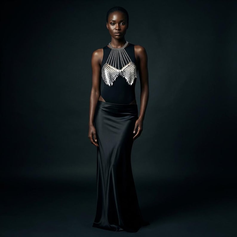 May include: A woman wearing a black bodysuit with a silver chainmail top and a long black satin skirt. The chainmail top has a unique design, and the skirt flows to the floor. The background is a dark, solid color.