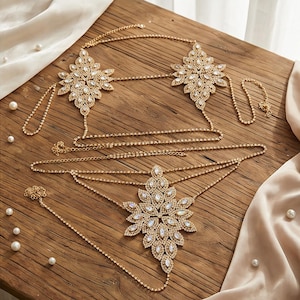 Peut inclure: Bijoux de corps dorés avec des accents de strass. Les bijoux présentent trois grands motifs floraux ornés reliés par de fines chaînes. Les pièces sont disposées sur une surface en bois avec des perles et du tissu drapé.