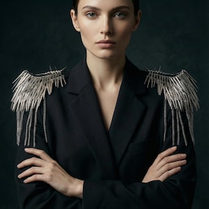 May include: A black blazer adorned with silver shoulder pieces. The shoulder embellishments have a spiky design with hanging chains. The model is wearing the blazer with arms crossed, creating a bold and edgy aesthetic.