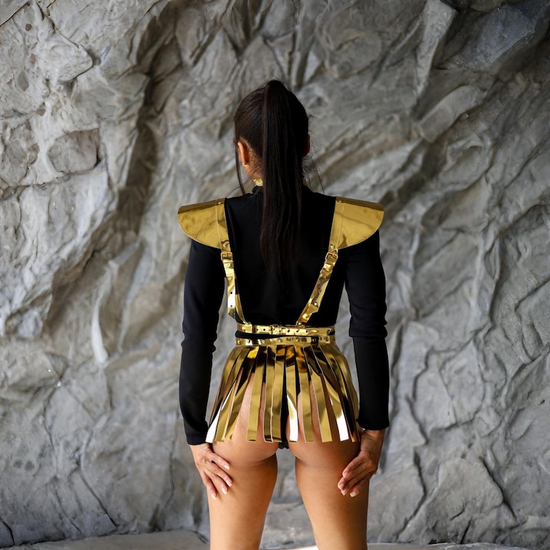 May include: A black long-sleeved top with gold metallic shoulder pads, suspenders, and a fringed skirt. The outfit is accessorized with a gold belt and is set against a textured gray stone wall.