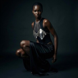 May include: A woman in a black dress and silver chainmail top. The dress has a thigh-high slit. The silver top is made of multiple silver discs and chains. The woman is crouching on a dark background.