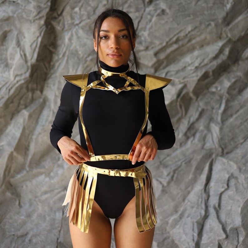 May include: A gold-colored metal harness and skirt set. The harness features shoulder accents and a geometric chest design. The skirt has vertical fringe. The set is worn over a black bodysuit. The background is a textured gray surface.
