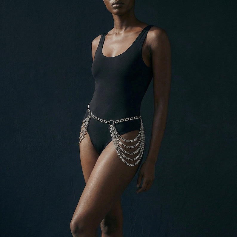 Op de afbeelding: Een zwarte bodysuit met een zilveren kettingriem. De kettingriem heeft een cirkelvormige sluiting en meerdere strengen die over de voorkant van de benen hangen. De bodysuit heeft een ronde halslijn en is mouwloos.
