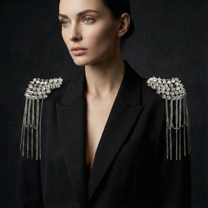 Pode incluir: Um blazer preto com enfeites de ombro prateados. Os enfeites apresentam aglomerados de pedras brilhantes e correntes penduradas, adicionando um toque de glamour ao casaco clássico. O blazer tem lapelas entalhadas e um corte sob medida.