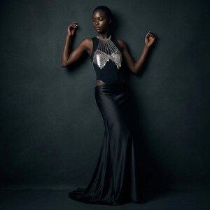 May include: A black sleeveless top with a silver chainmail design and a long black satin skirt. The model is standing against a dark gray background. The top has a high neckline and a cutout on the side.