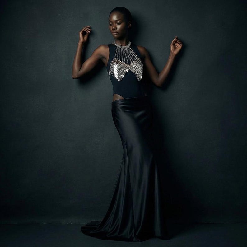 May include: A black sleeveless top with a silver chainmail design and a long black satin skirt. The model is standing against a dark gray background. The top has a high neckline and a cutout on the side.