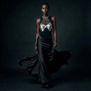 May include: A black sleeveless bodysuit with a silver chainmail design on the chest, paired with a flowing black satin skirt. The model is walking forward against a dark background, with the skirt billowing.