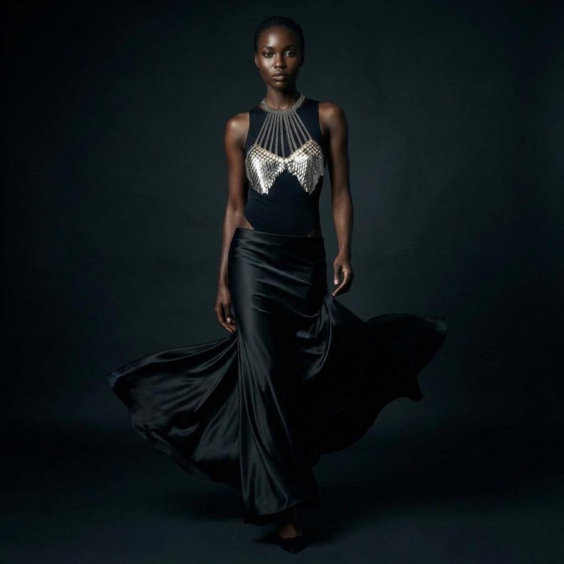 May include: A black sleeveless bodysuit with a silver chainmail design on the chest, paired with a flowing black satin skirt. The model is walking forward against a dark background, with the skirt billowing.