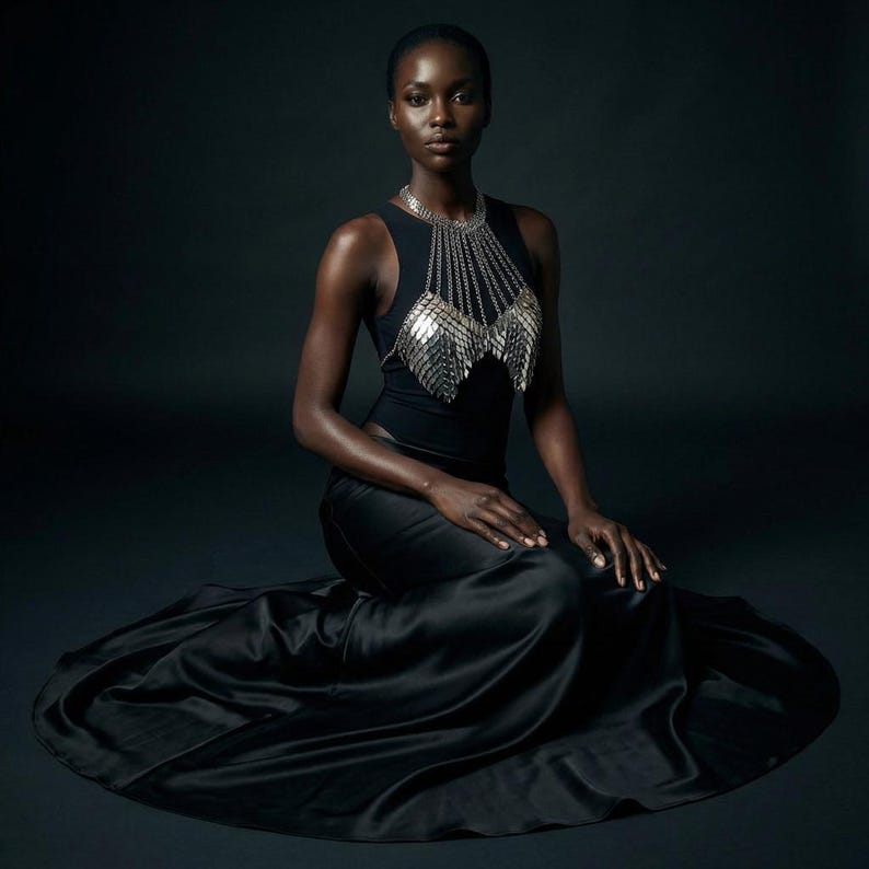May include: A woman wearing a black sleeveless top with a silver chainmail chest piece and a matching necklace. She is also wearing a long, flowing black satin skirt. The background is a dark gray.