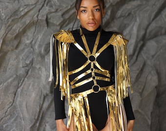 May include: A black bodysuit with gold-colored harness detailing and fringe. The harness features a series of connected rings and straps. The fringe is attached to the shoulders and waist, creating a layered effect. The background is a textured gray wall.