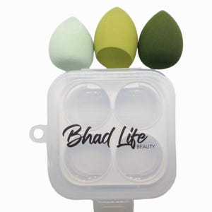 May include: Three teardrop-shaped makeup sponges in shades of green sit above a clear plastic case. The case has four circular compartments and the words "Bhad Life BEAUTY" in black script.