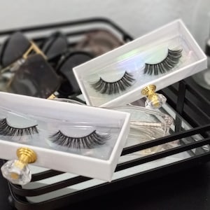 May include: A black metal tray with a mirrored bottom holds two white boxes of false eyelashes. The boxes have gold and crystal knobs. A black box with the text "Cloud Lite" is also visible, along with sunglasses and a perfume bottle.