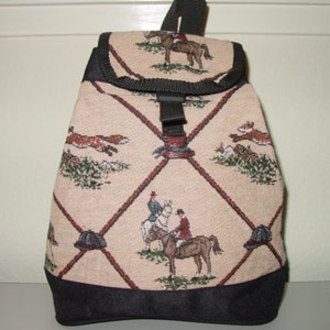 May include: A beige backpack with a black base and accents. The bag features a repeating pattern of riders on horseback, foxes, and hunting hats. The design is framed by a dark red diamond pattern. The bag has a top handle and adjustable straps.