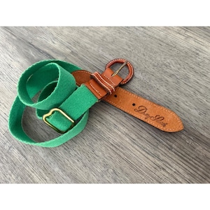 Puede incluir: Un cinturón de lona verde con un extremo de cuero marrón y una hebilla de metal color latón. La parte de cuero tiene grabado el texto Dotty Smith en cursiva. El cinturón incluye una trabilla de lona verde y un ajustador de metal.