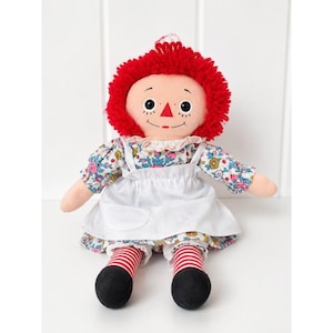 May include: A fabric doll with bright red yarn hair, black button eyes, and a stitched triangular nose. The doll wears a floral print dress with blue, pink, and yellow flowers, a white apron, and red and white striped stockings with black feet.