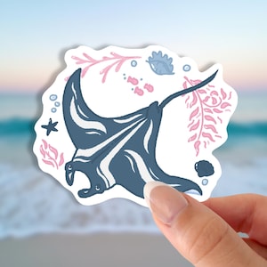 May include: A sticker featuring a stylized illustration of a manta ray in shades of blue and white, surrounded by pink coral, a starfish, and seashells. The sticker has a white border and is held by a hand.