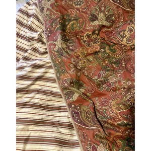 May include: A bedspread with a paisley pattern in shades of red, green, and gold. The bedspread is next to a striped sheet in brown and white. The bedding is folded and appears to be made of a soft fabric.