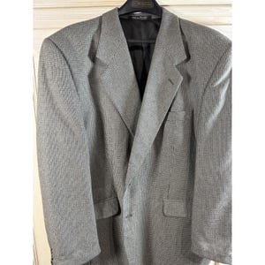 May include: A grey blazer with a subtle chequered pattern. The blazer features a notched lapel, two front pockets, and a single-button closure. The interior lining is black.