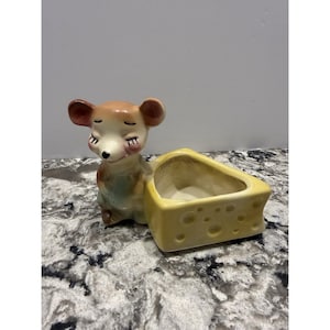 May include: A ceramic figurine of a mouse with closed eyes, wearing a light green outfit, next to a yellow cheese-shaped container. The mouse has brown ears and a pink nose. The cheese container has holes.