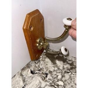May include: A wooden wall hook with a light brown finish. The hook has two curved metal arms with white ceramic knobs. The hook is attached to a wooden base with brass hardware.