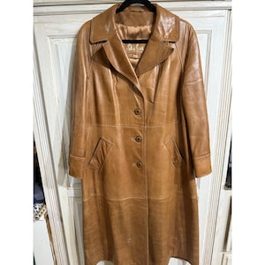 以下が含まれることがあります： ノッチカラー、ボタン留め、2つのフロントポケットが付いた、ヴィンテージの膝丈タンレザージャケット。 ジャケットは黒いハンガーに吊るされています。 ジャケットには「She's Got It」と書かれたラベルが付いています。