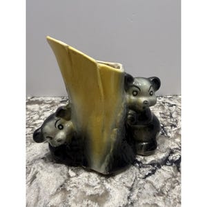 Puede incluir: Un jarrón de cerámica vintage con un diseño de tronco de árbol con dos cachorros de oso asomándose. El jarrón tiene una combinación de colores amarillo y negro, con los osos en un acabado brillante. Una pieza decorativa encantadora.
