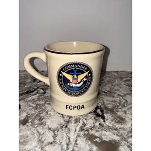 May include: A cream-colored ceramic mug with a gold-toned rim and a handle on the left side. The front features a circular emblem with an eagle, anchor, and the text COMMANDER U.S. NAVAL FORCES JAPAN. The letters FCPOA are printed in black below the emblem.