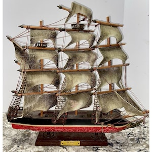 May include: A model ship named Sea Witch Clipper 1846 with a black and red hull featuring a cracked paint finish. The vessel has three wooden masts, multiple tan sails with dark edges, and intricate rigging lines attached to the masts and deck.