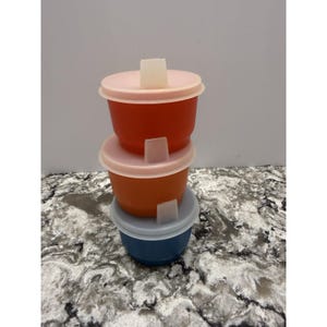 May include: Stack of three small, colorful plastic containers with lids. The containers are red, orange, and blue, with light pink and white lids. Each lid has a small, rectangular tab.