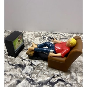 Puede incluir: Una figura en miniatura representa a una persona relajándose en un sillón marrón, sosteniendo un mando a distancia y una bebida. La persona lleva una camisa roja y pantalones azules. Un televisor negro muestra un partido de fútbol americano.