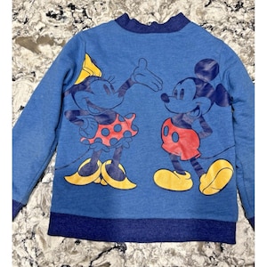 以下が含まれることがあります： ミッキーとミニーマウスが描かれた青いスウェットシャツ。ミニーは赤い水玉模様のスカートと黄色の靴を履いています。ミッキーは赤いショーツと黄色の靴を履いています。スウェットシャツは青い襟と袖口が付いています。