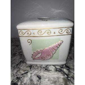 May include: A white ceramic container with a lid, featuring gold scrollwork and a pink seashell design on a light green background. The container has a square shape with rounded edges, suitable for bathroom or kitchen decor.