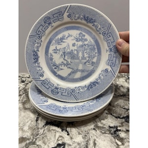 May include: A stack of four white ceramic plates with a blue and white Chinoiserie design. The plates feature a central landscape scene and a decorative border. The plates are stacked on top of each other.