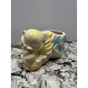 May include: A vintage ceramic planter in the shape of a bear cub. The planter is painted in pastel yellow, blue, and cream colors. The bear cub is curled up, with its head resting on its paws. The planter has a rectangular opening for plants.