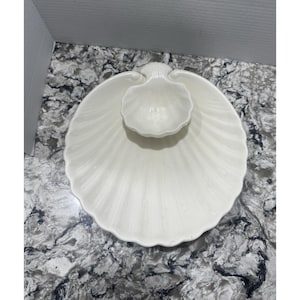 May include: A white ceramic serving dish set in a seashell design. The set includes a large, scalloped-edged platter and a smaller, shell-shaped bowl. The dish has a glossy finish and is suitable for serving appetizers or snacks.