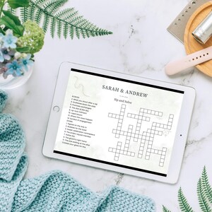 Op de afbeelding: Een tablet met een aangepaste kruiswoordpuzzel getiteld Sip and Solve voor Sarah en Andrew. Het scherm toont genummerde aanwijzingen en een raster van lege vakjes. De tablet is omringd door een lichtblauwe gebreide deken en een vaas met bloemen.