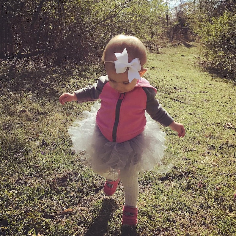 Pode incluir: Uma menina pequena vestindo uma jaqueta rosa e uma saia tutu branca com um la&ccedil;o branco no cabelo. Ela est&aacute; caminhando por um caminho gramado.