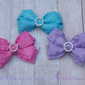 May include: Three fabric hair bows with ruffled edges and a rhinestone center. The bows are in shades of pink, blue, and purple.