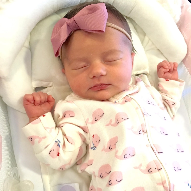 May include: A sleeping baby wearing a white onesie with a pink whale pattern and a pink bow headband.