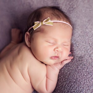 May include: A newborn baby girl sleeping on a purple blanket, wearing a pink and gold headband with a bow.